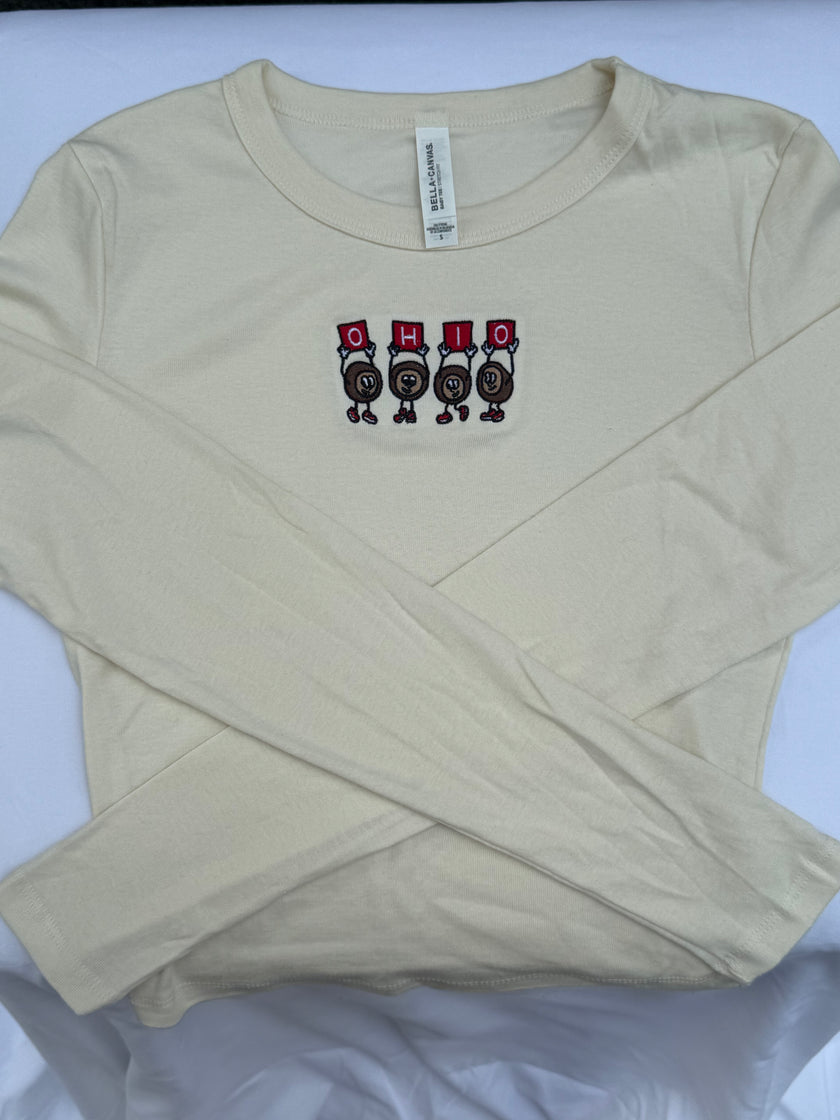 A beige long-sleeve t-shirt with a graphic of four cartoon characters wearing red and black hats spelling 'OHIO' on the chest, laid flat on a white background.