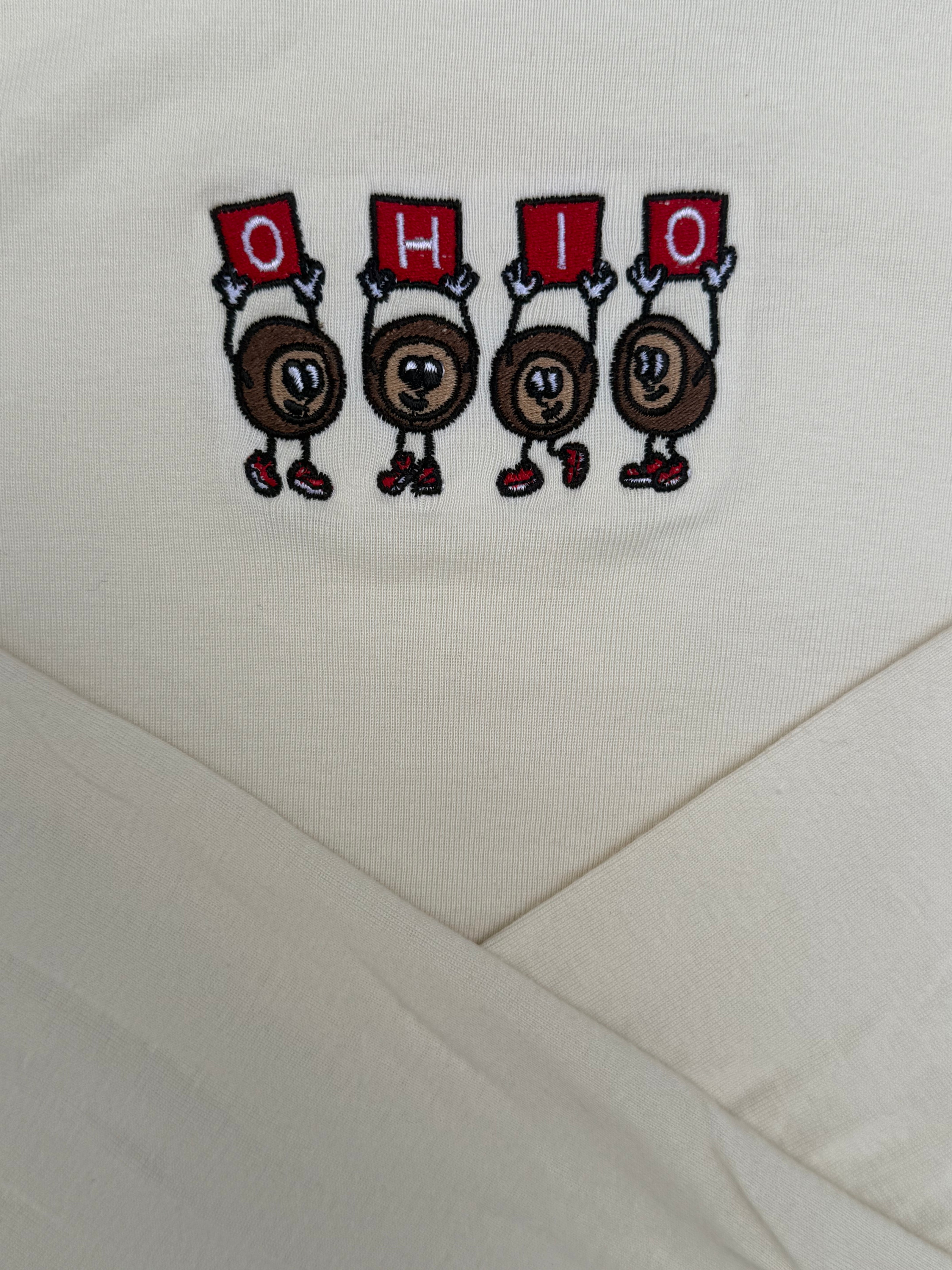 Close-up of a cream-colored fabric garment featuring an embroidered graphic of four cartoon characters holding red signs that spell 'OHIO'.