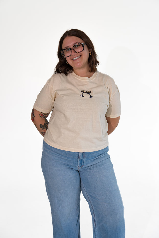 Smiling woman wearing glasses and standing against a white background, wearing a cream-colored short-sleeve graphic tee with an espresso martini patch, paired with light-wash blue jeans.