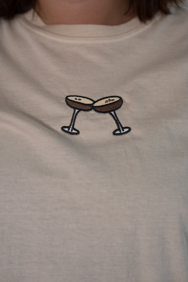 Close-up of the front of a cream-colored t-shirt featuring a detailed embroidered graphic of two espresso martini glasses clinking together.