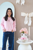 Model wearing a light pink Fiancé bow crewneck sweatshirt with dark blue jeans in a bridal-inspired setting.