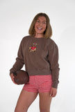 A woman modeling a brown crewneck sweatshirt with a teddy bear graphic, paired with red and white gingham shorts, holding a brown football against a white studio background.