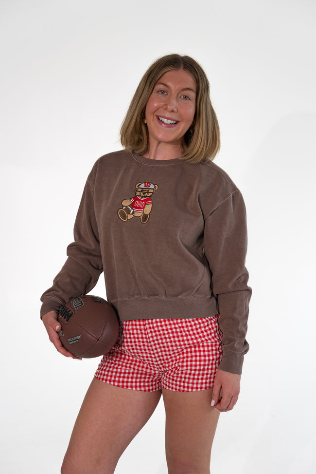 A woman modeling a brown crewneck sweatshirt with a teddy bear graphic, paired with red and white gingham shorts, holding a brown football against a white studio background.