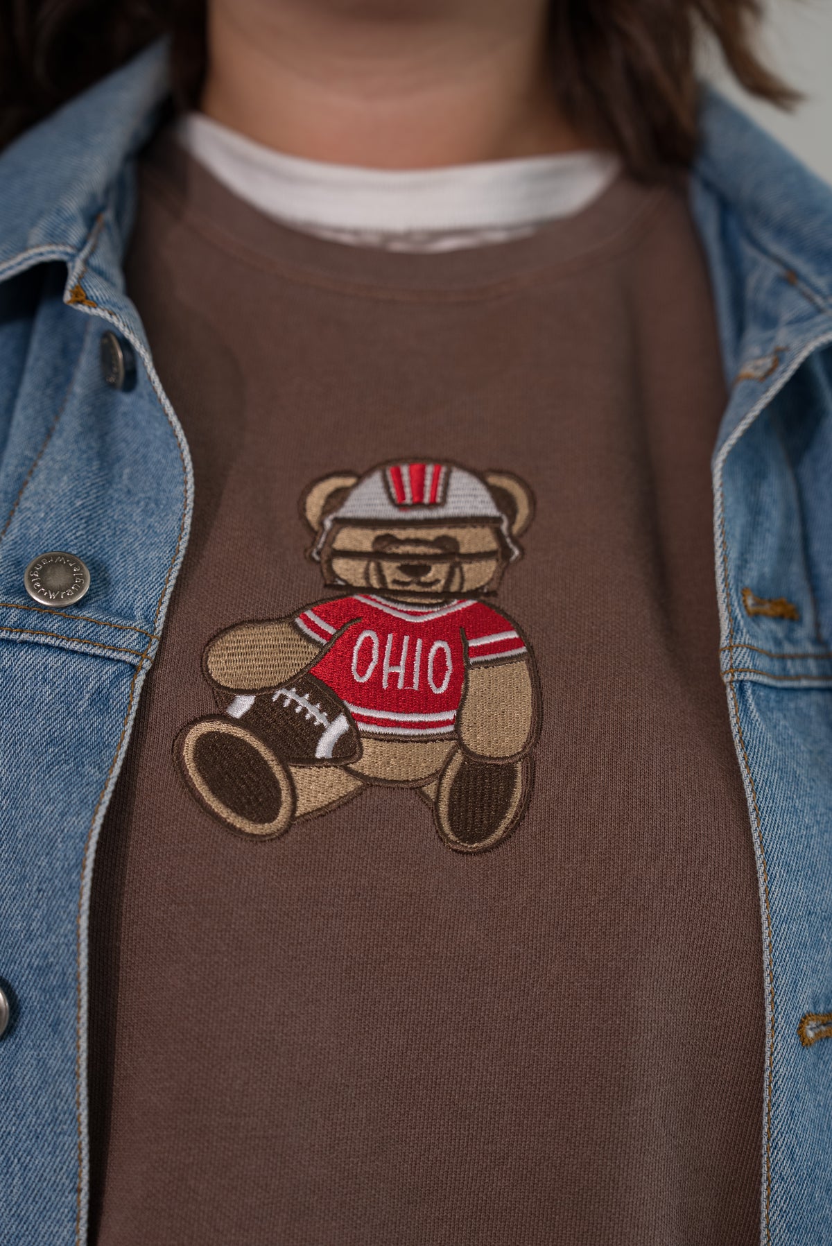 A close-up of a person wearing a brown crewneck sweatshirt with a teddy bear graphic wearing a red and white Ohio football jersey, styled under a light blue denim jacket.