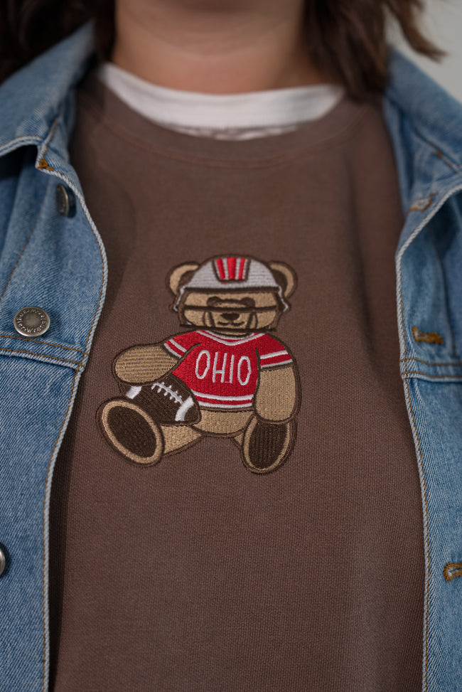 A close-up of a person wearing a brown crewneck sweatshirt with a teddy bear graphic wearing a red and white Ohio football jersey, styled under a light blue denim jacket.