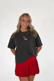 Smiling woman wearing a charcoal gray short-sleeve t-shirt with a small embroidered dog patch on the chest, paired with a bright red pleated skirt.