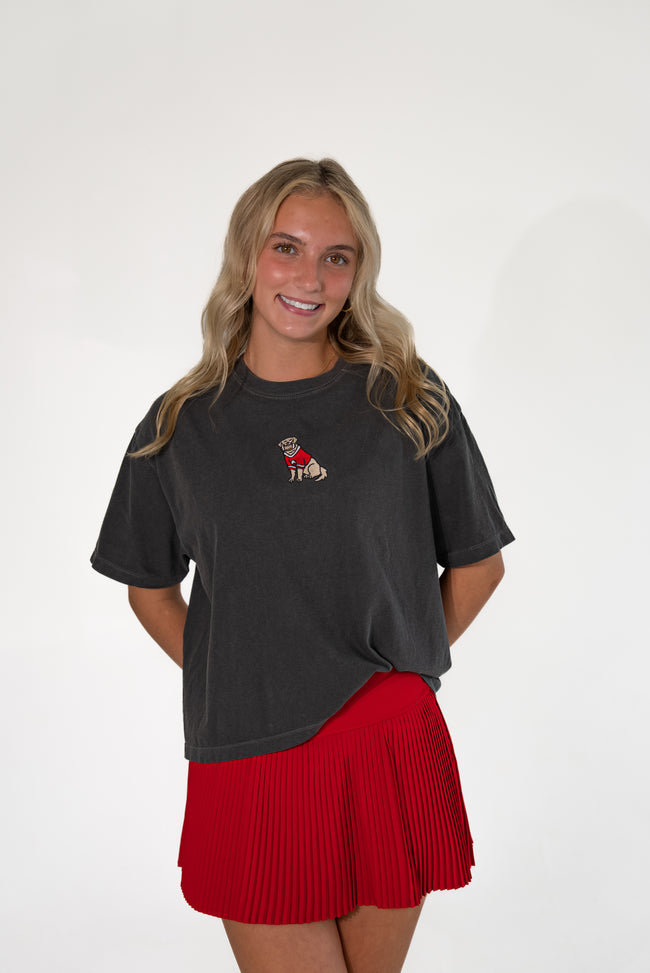 Smiling woman wearing a charcoal gray short-sleeve t-shirt with a small embroidered dog patch on the chest, paired with a bright red pleated skirt.