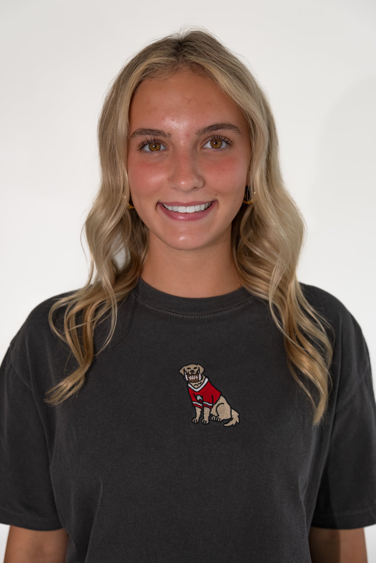 A smiling blonde woman wearing a faded black t-shirt with a small embroidered graphic of a dog in a red sweater on the left chest, set against a plain white background.