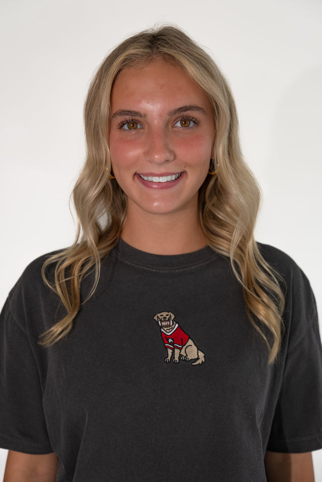 A smiling blonde woman wearing a faded black t-shirt with a small embroidered graphic of a dog in a red sweater on the left chest, set against a plain white background.