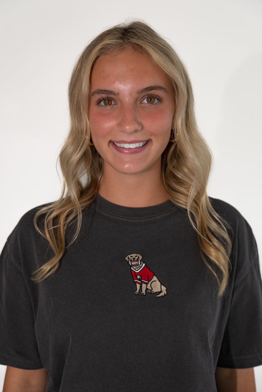 A smiling blonde woman wearing a faded black t-shirt with a small embroidered graphic of a dog in a red sweater on the left chest, set against a plain white background.