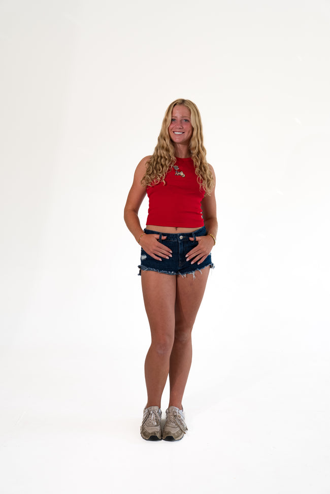 Smiling blonde woman standing against a white background, wearing a bright red ribbed high-neck tank top with an embroidered dog patch, dark wash distressed denim cutoff shorts, and sneakers