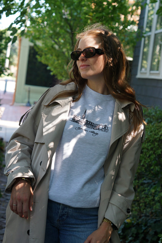 Woman wearing a heather gray German Village embroidered crewneck layered under a beige trench coat.