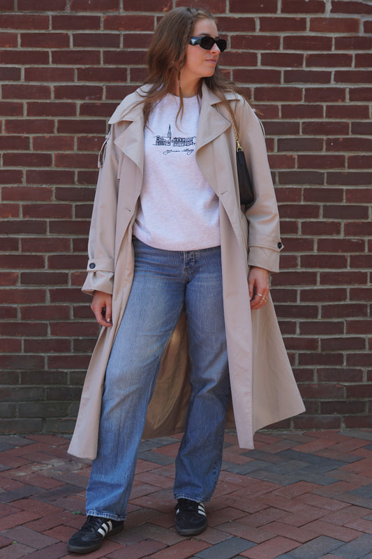Full-body view of a woman styling a German Village embroidered crewneck sweatshirt with a beige trench coat and blue jeans.