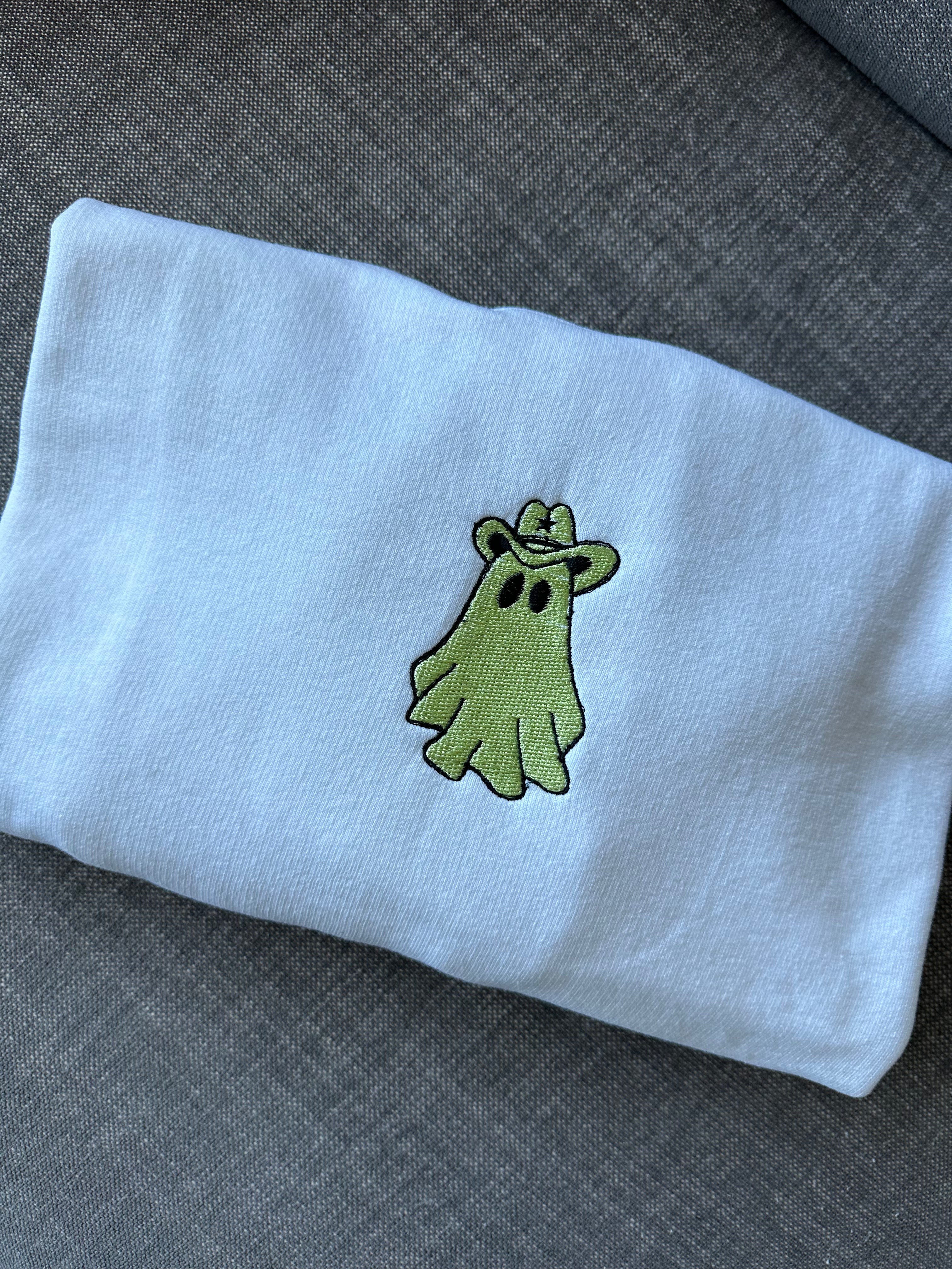 Close-up of a white crewneck sweatshirt featuring an embroidered green ghost wearing a cowboy hat, centered on the chest with black outline detailing.