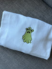 Close-up of a white crewneck sweatshirt featuring an embroidered green ghost wearing a cowboy hat, centered on the chest with black outline detailing.