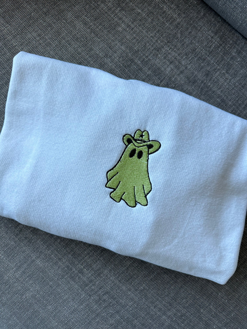 Close-up of a white crewneck sweatshirt featuring an embroidered green ghost wearing a cowboy hat, centered on the chest with black outline detailing.
