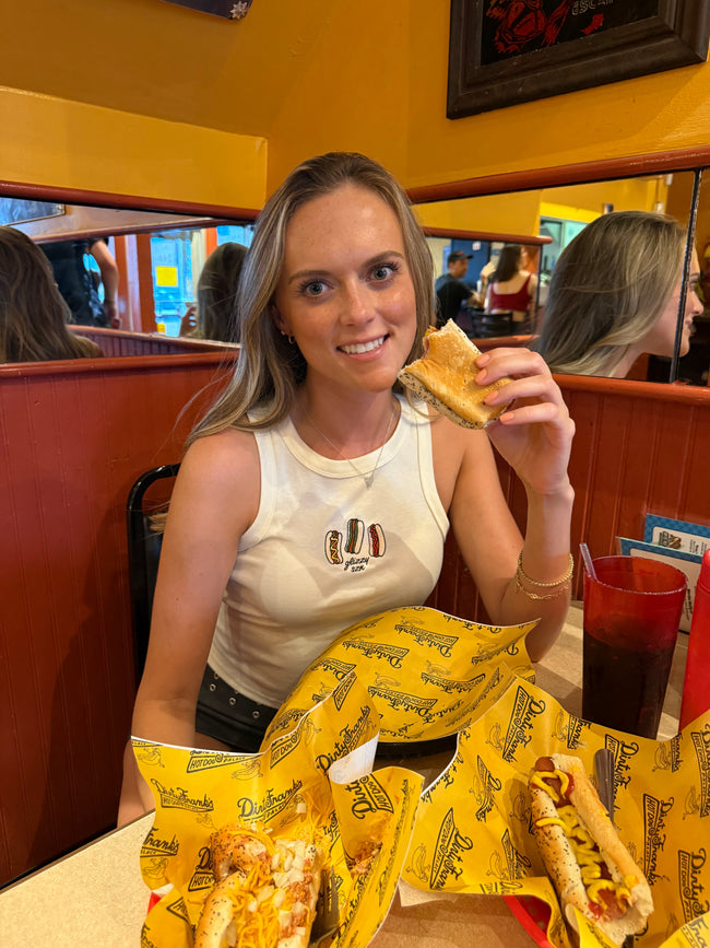 Model wearing the Glizzy Season embroidered tank while enjoying hot dogs at a restaurant.