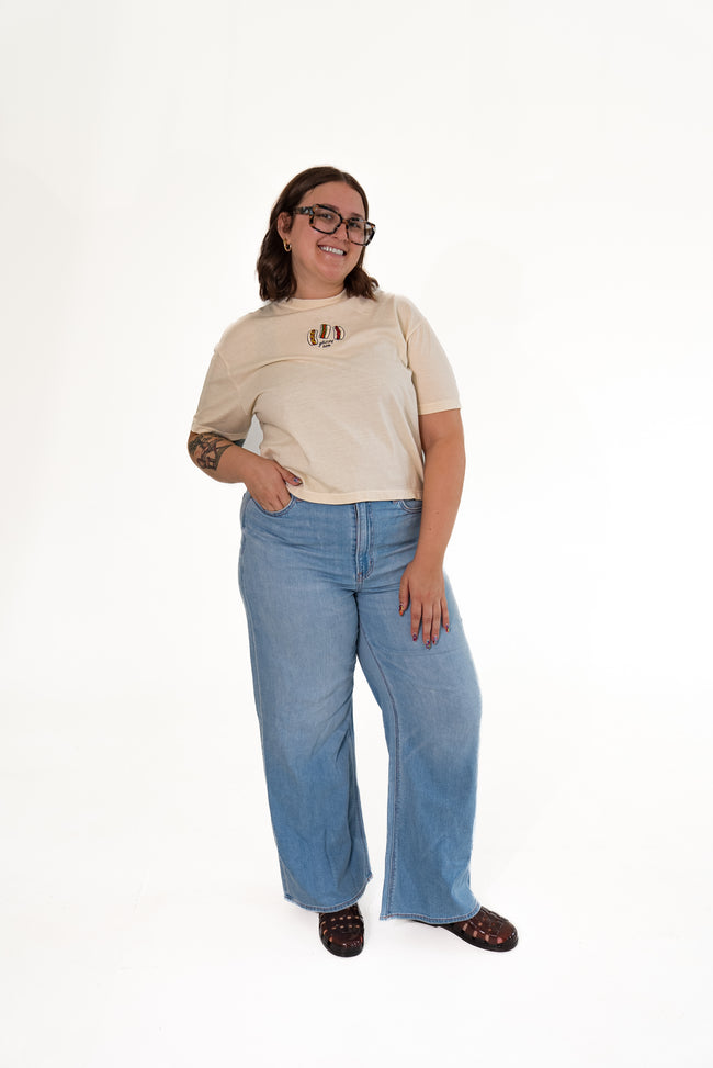Smiling woman wearing glasses and standing against a white background, wearing a cream-colored short-sleeve graphic tee with a hot dog patch, paired with light-wash blue wide-leg jeans and dark sandals.