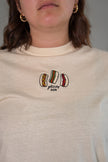 Close-up of the front of a cream-colored t-shirt featuring a detailed embroidered graphic of three hot dogs with various toppings and the text 