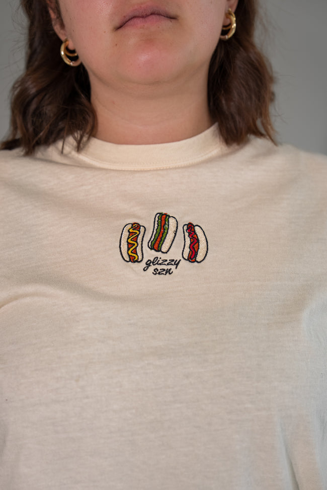 Close-up of the front of a cream-colored t-shirt featuring a detailed embroidered graphic of three hot dogs with various toppings and the text 
