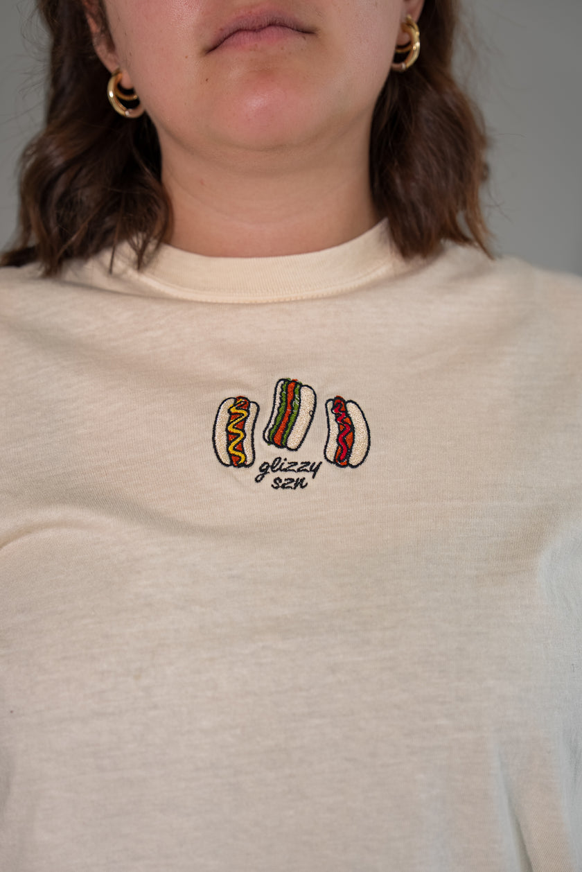 Close-up of the front of a cream-colored t-shirt featuring a detailed embroidered graphic of three hot dogs with various toppings and the text 