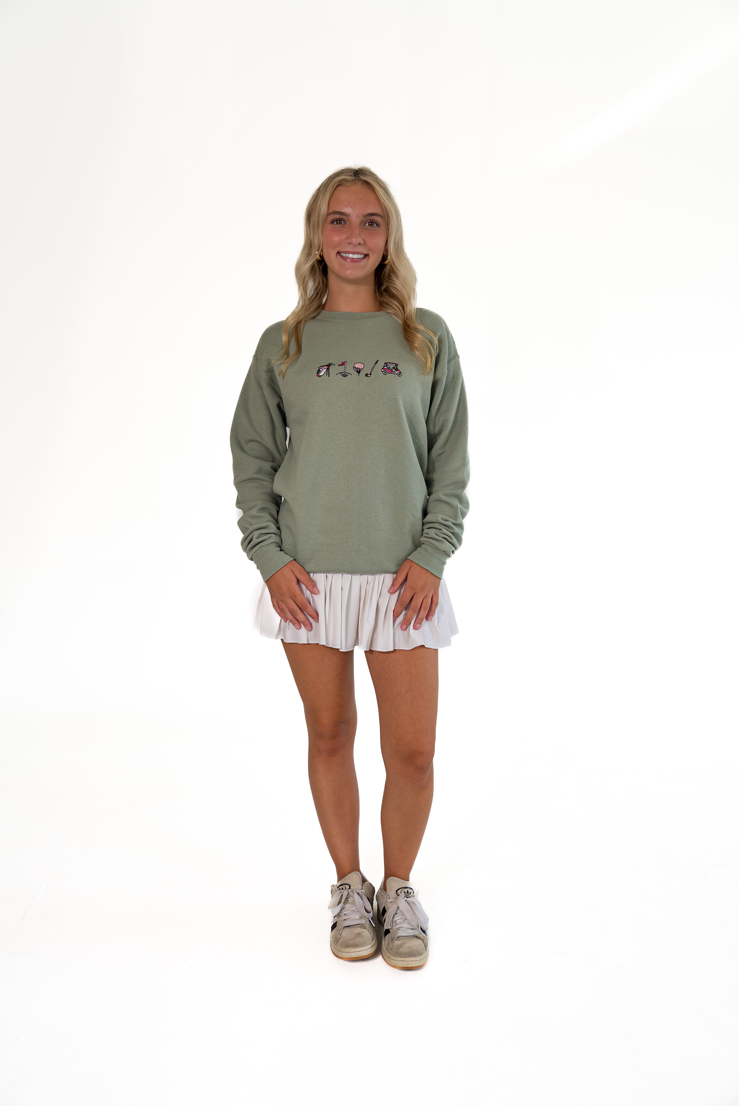 A young woman stands against a seamless white studio background wearing a sage green crewneck sweatshirt with a small graphic on the chest, a white pleated mini skirt, and gray sneakers.