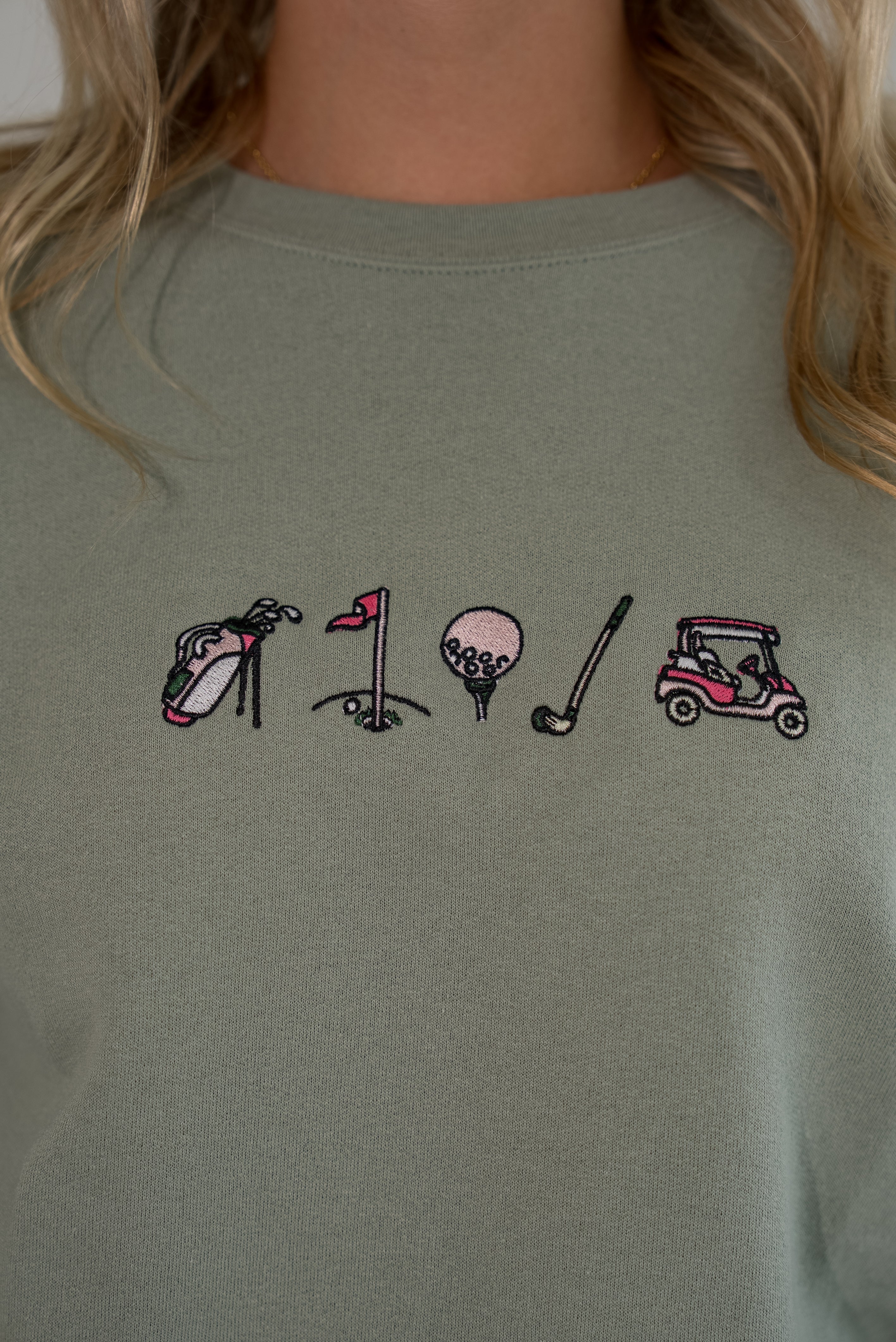 Close-up of a person wearing a muted sage green sweatshirt with a small embroidered golf-themed graphic across the chest, including a golf bag, flag, ball, club, and cart.