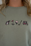 Close-up of a person wearing a muted sage green sweatshirt with a small embroidered golf-themed graphic across the chest, including a golf bag, flag, ball, club, and cart.