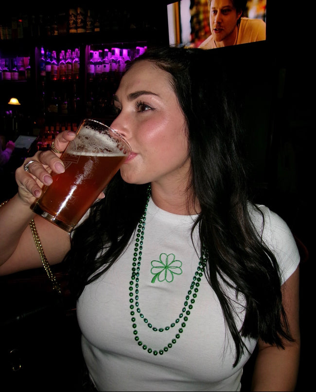 Woman wearing a white fitted baby tee with green embroidered four-leaf clover design.