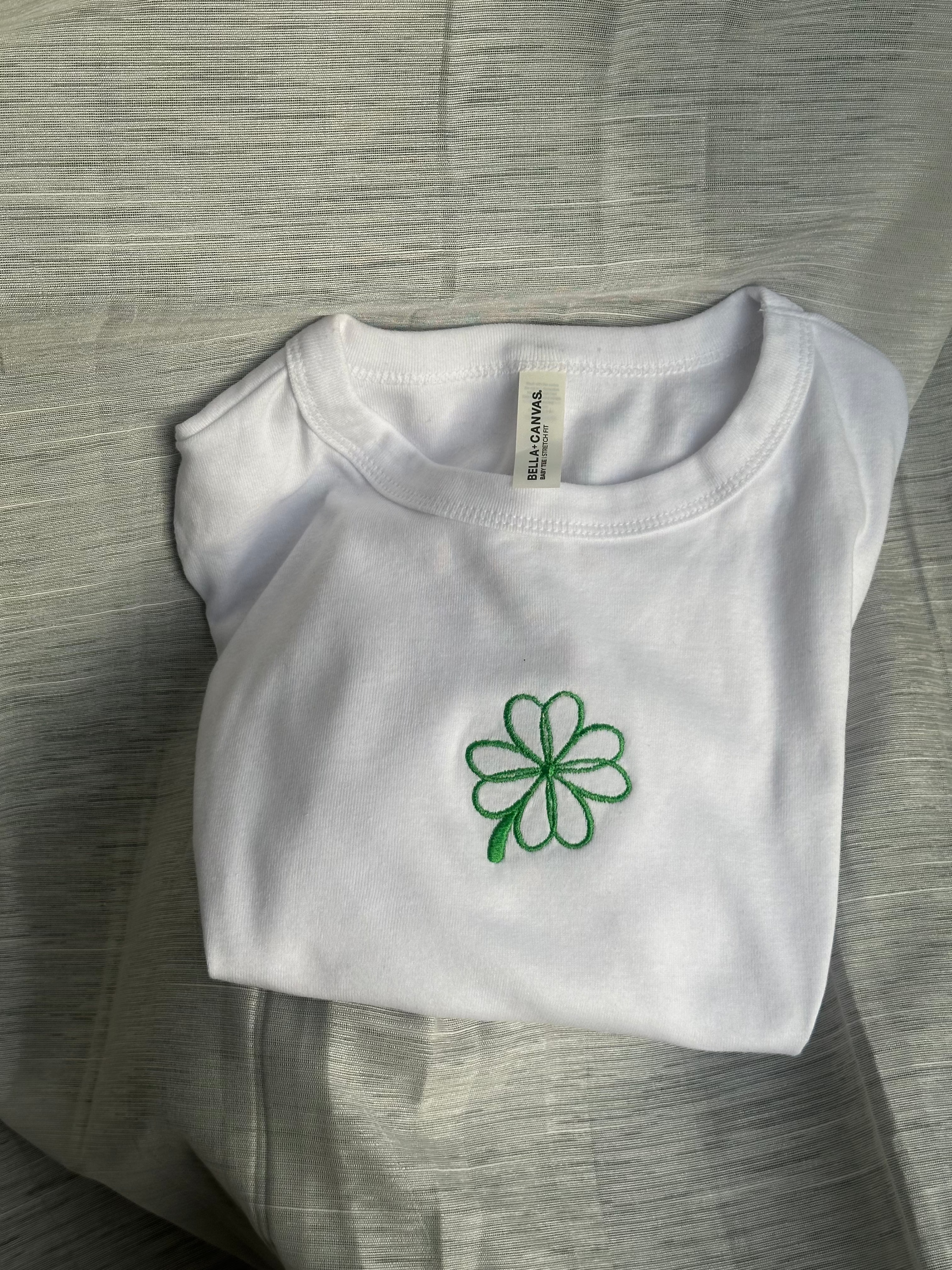 White fitted baby tee with green embroidered four-leaf clover design.