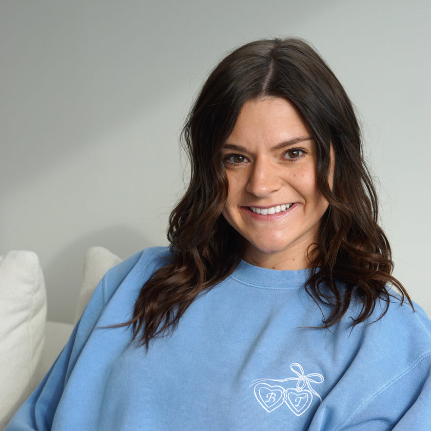 Model wearing a light blue crewneck sweatshirt embroidered with personalized heart locket initials.