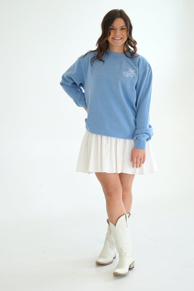 Model wearing a light blue embroidered Heart Locket Crew sweatshirt with a white pleated skirt and white cowboy boots.