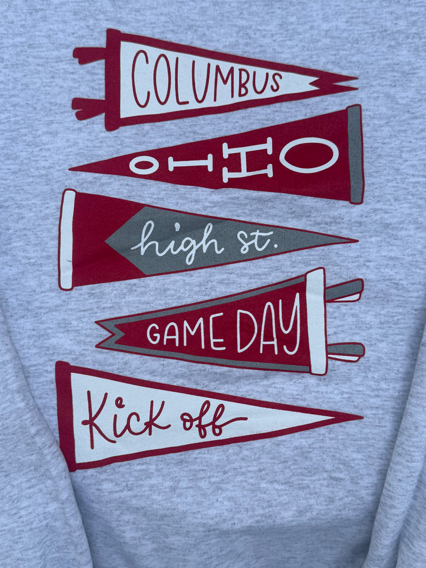 Close-up of a heather gray sweatshirt featuring a graphic print of five overlapping sports pennants in maroon and gray with white lettering that reads 'COLUMBUS', 'OHIO', 'high st.', 'GAME DAY', and 'Kick off'.
