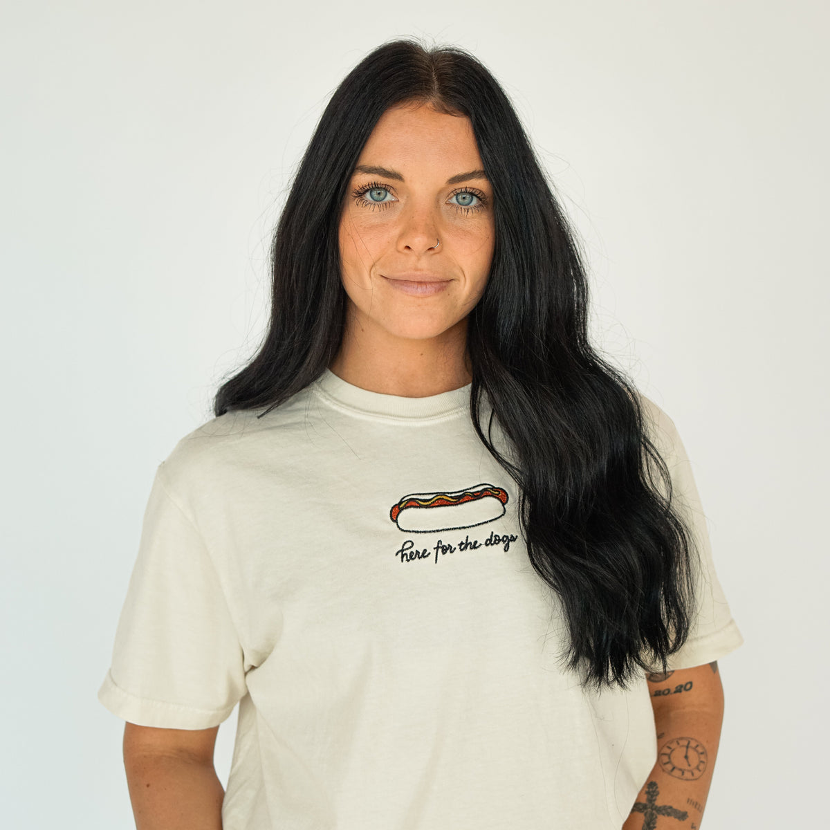 Woman wearing a cream embroidered T-shirt featuring a hot dog graphic and “Here for the dogs” lettering, styled for a casual baseball game day look.
