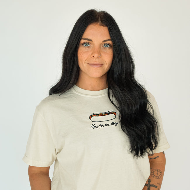 Woman wearing a cream embroidered T-shirt featuring a hot dog graphic and “Here for the dogs” lettering, styled for a casual baseball game day look.