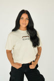 Woman wearing a cream embroidered T-shirt featuring a hot dog graphic and “Here for the dogs” lettering, styled for a casual baseball game day look.