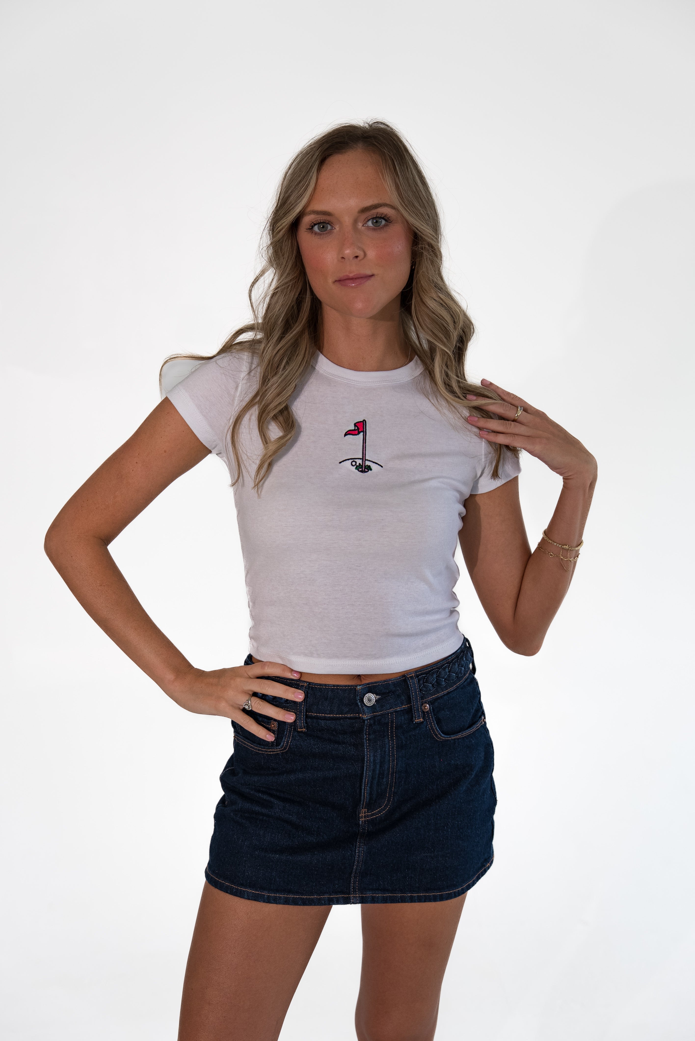 A woman modeling a white short-sleeve crop top with a small black and red golf flag graphic on the chest, paired with a dark denim mini skirt, against a plain white studio background.