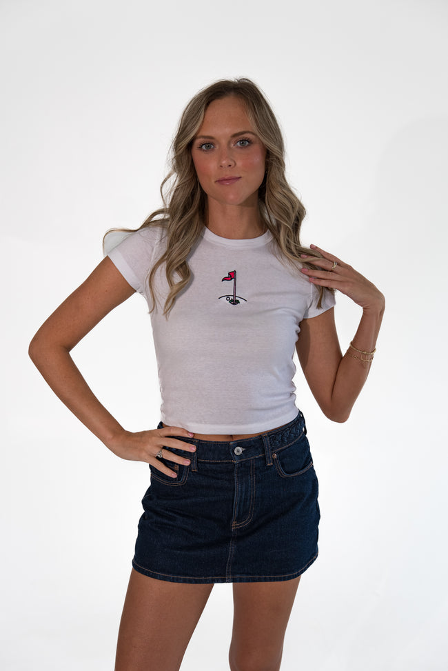 A woman modeling a white short-sleeve crop top with a small black and red golf flag graphic on the chest, paired with a dark denim mini skirt, against a plain white studio background.