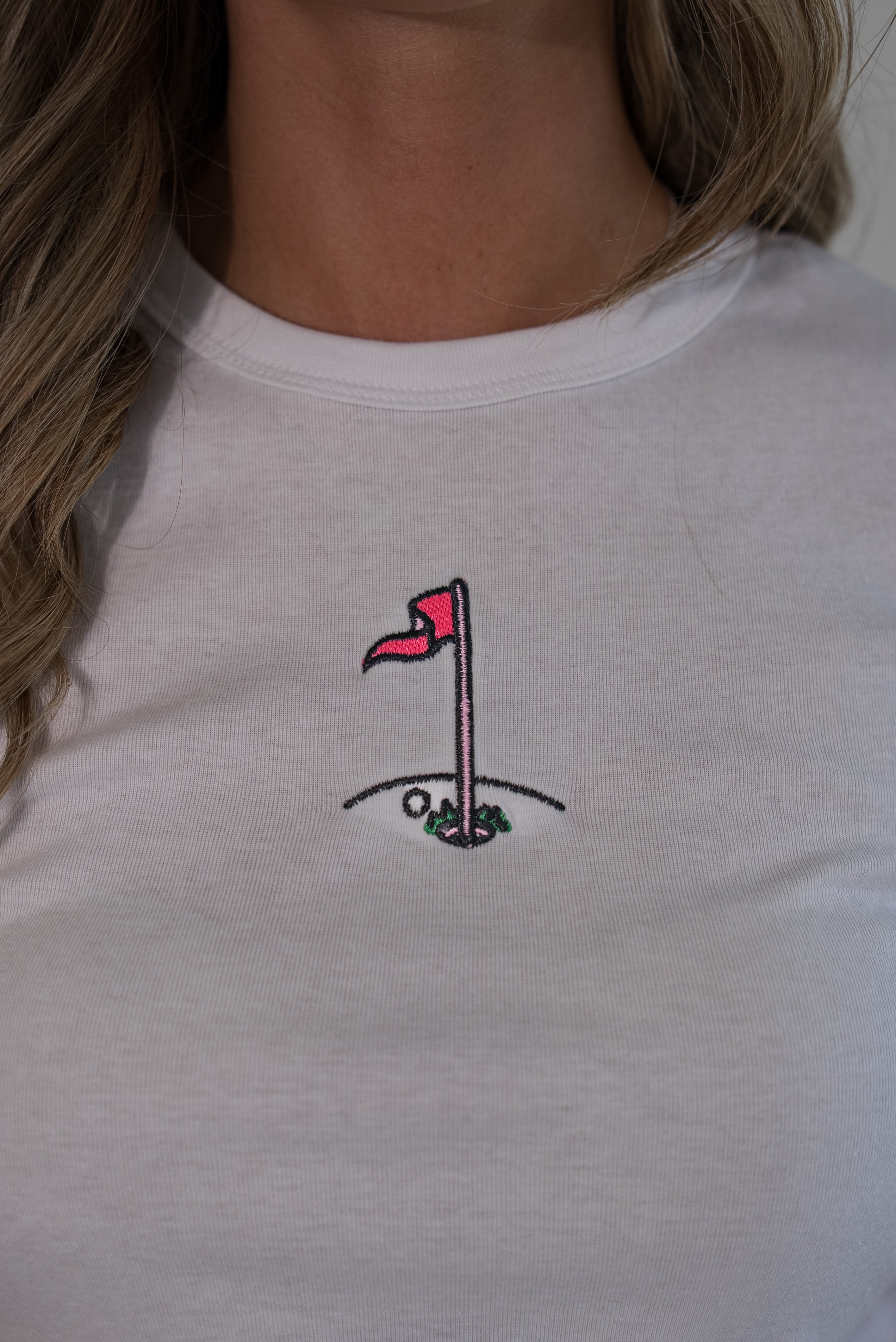 A close-up of a person wearing a white crewneck t-shirt with a small embroidered golf hole graphic on the chest.