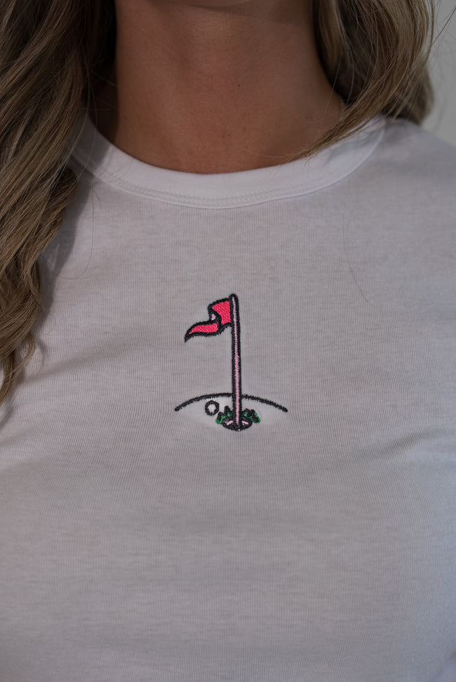 A close-up of a person wearing a white crewneck t-shirt with a small embroidered golf hole graphic on the chest.
