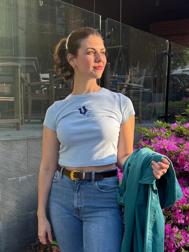 Woman wearing a light blue fitted baby tee with a small navy embroidered horseshoe, paired with high-waisted jeans and a belt outdoors.