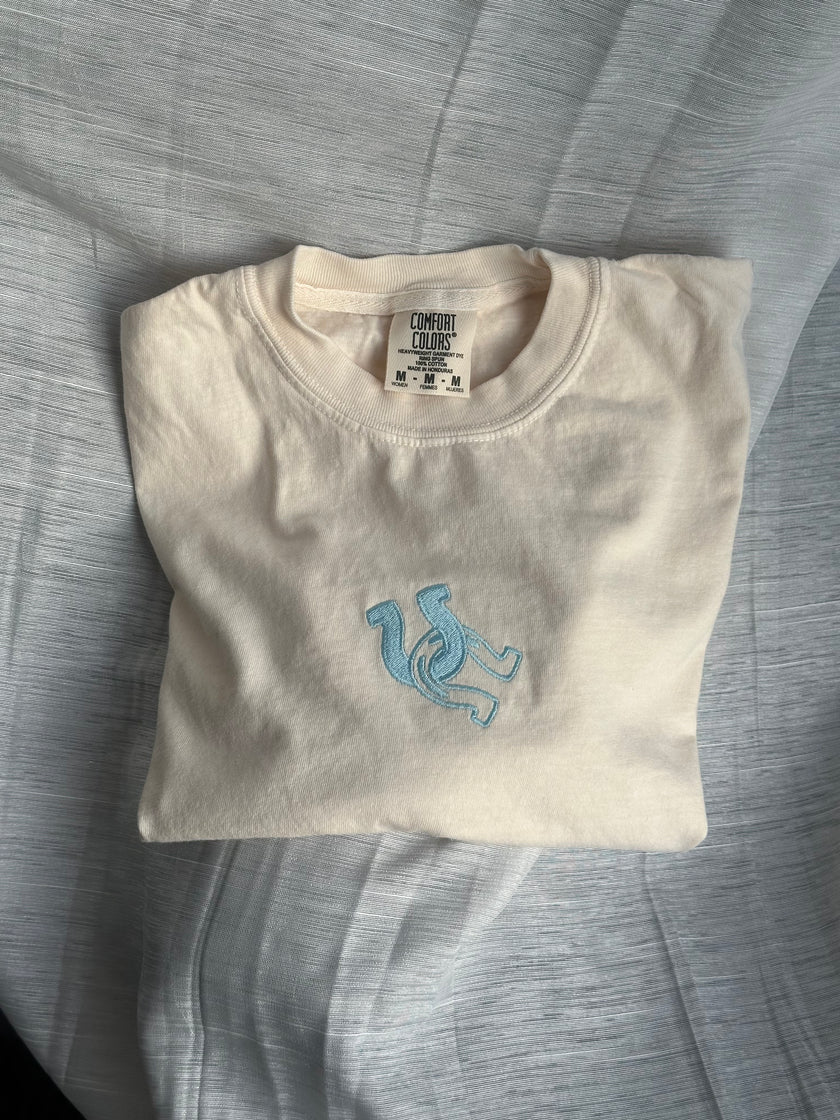 Ivory Comfort Colors boxy tee with a light blue embroidered horseshoe design, folded on a fabric surface.