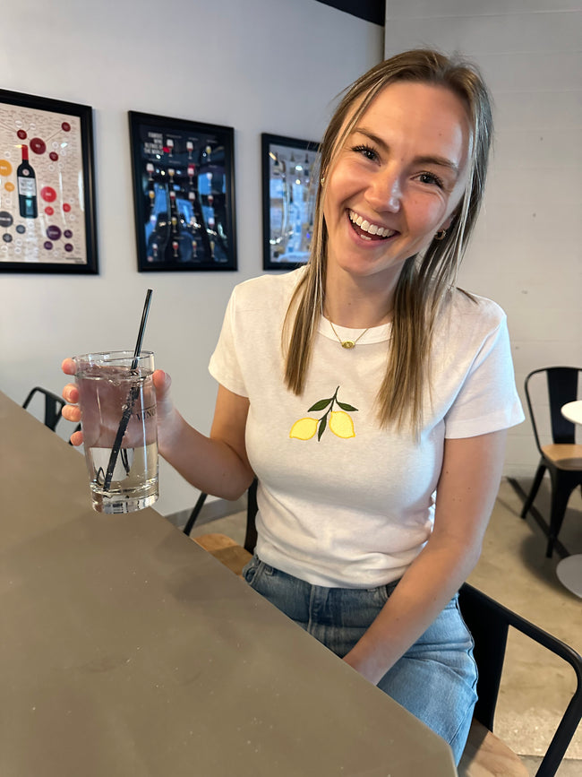 Model wearing the Lemon Embroidered Baby Tee with blue jeans while enjoying a drink at a café.