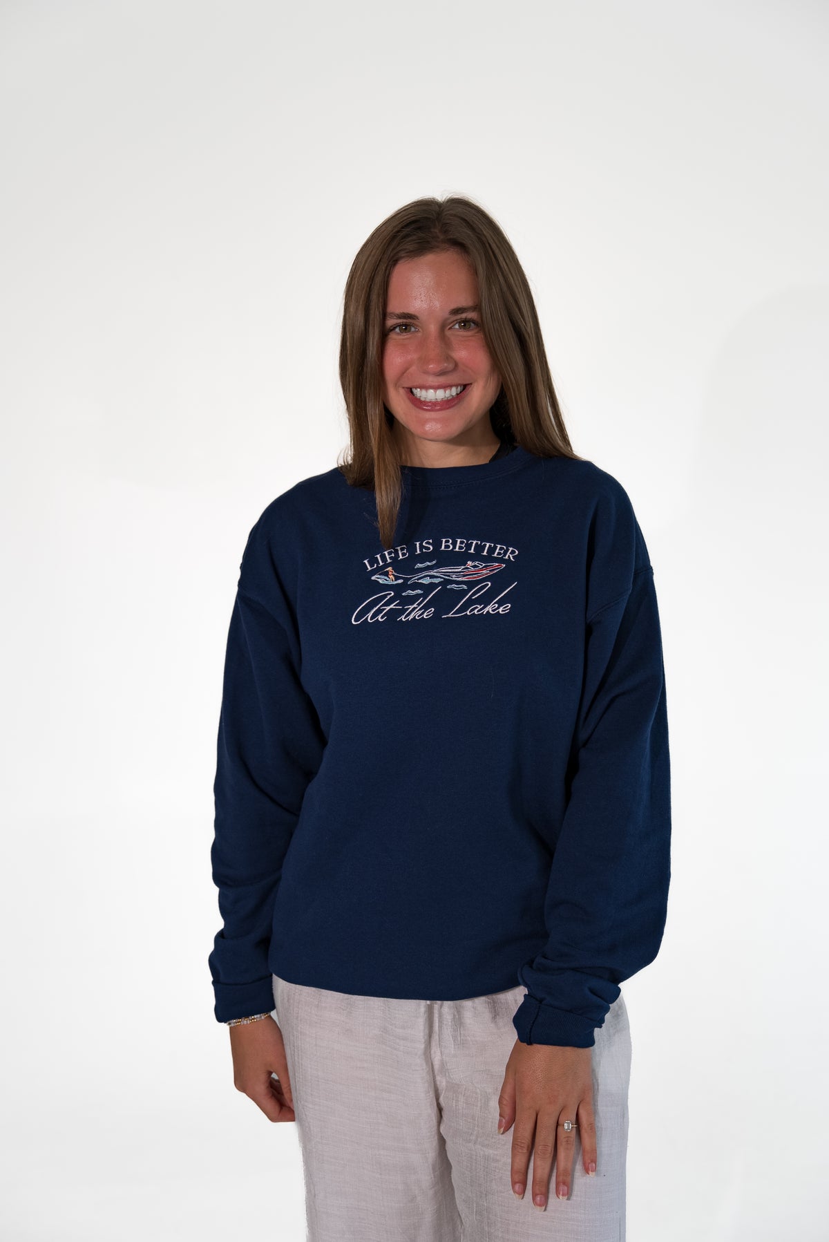 Smiling woman standing against a white background, wearing a navy blue crewneck sweatshirt with white and red embroidered text that reads 