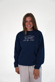 Smiling woman standing against a white background, wearing a navy blue crewneck sweatshirt with white and red embroidered text that reads 