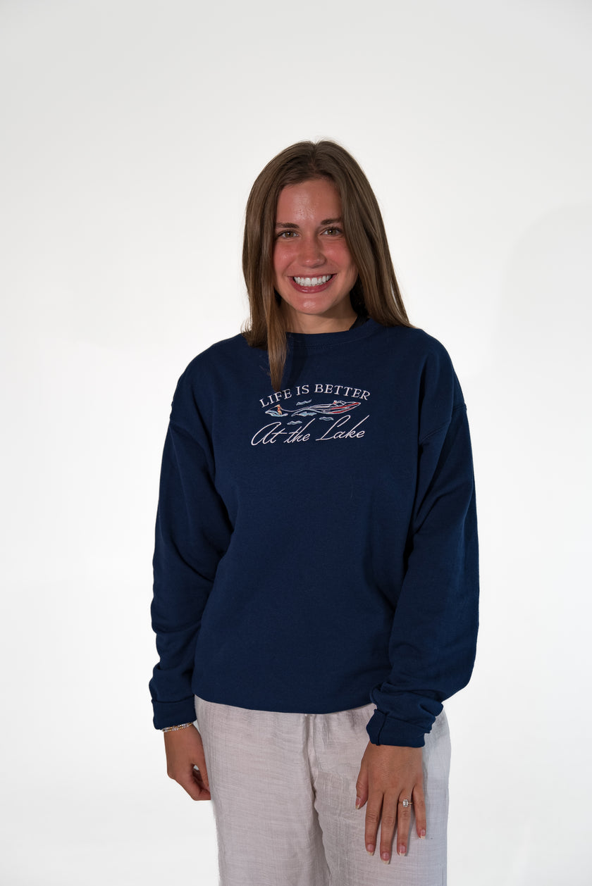 Smiling woman standing against a white background, wearing a navy blue crewneck sweatshirt with white and red embroidered text that reads 