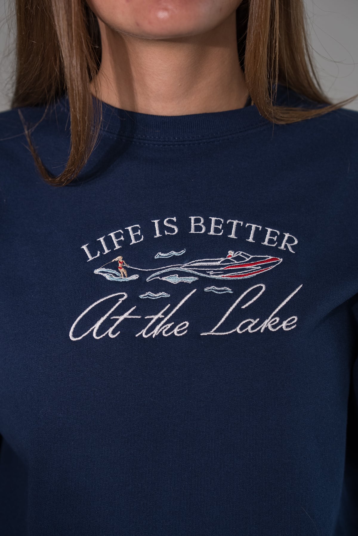 A close-up of a person wearing a navy blue crewneck sweatshirt with a graphic print that reads 'LIFE IS BETTER At the Lake' in white script and outline, featuring illustrations of a speedboat and a paddleboarder.