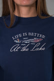 A close-up of a person wearing a navy blue crewneck sweatshirt with a graphic print that reads 'LIFE IS BETTER At the Lake' in white script and outline, featuring illustrations of a speedboat and a paddleboarder.