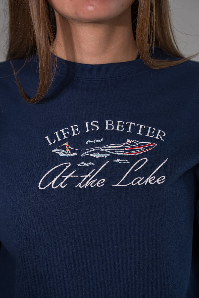 A close-up of a person wearing a navy blue crewneck sweatshirt with a graphic print that reads 'LIFE IS BETTER At the Lake' in white script and outline, featuring illustrations of a speedboat and a paddleboarder.