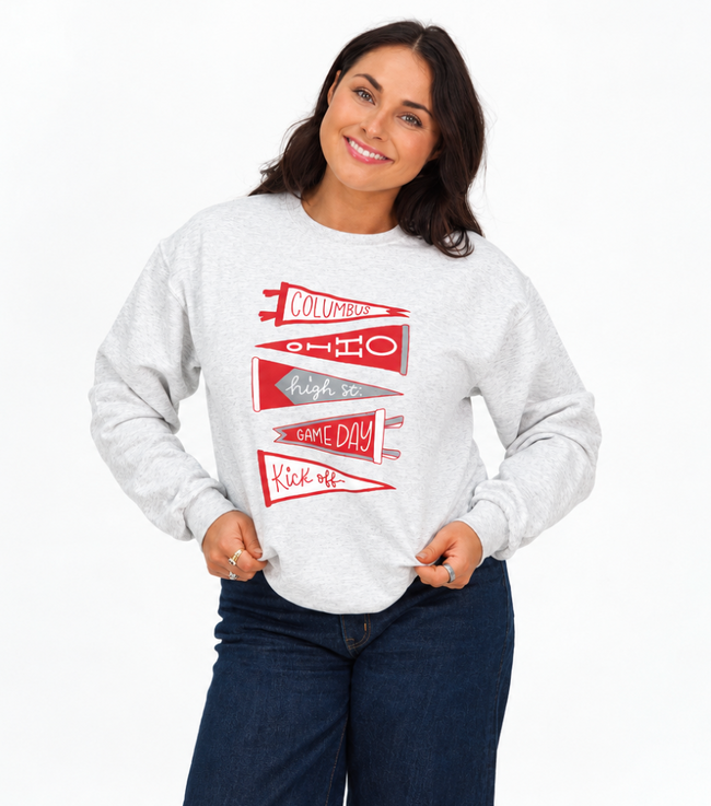 Woman wearing a white heather crewneck sweatshirt featuring red and white pennant graphics with “Columbus,” “Ohio,” “High St.,” “Game Day,” and “Kickoff” lettering, paired with dark blue jeans.